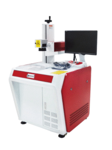 A red and white laser marking machine, viewed from a slightly elevated angle, is positioned against a clean white background. The machine has a substantial red base with a white front panel that includes a small door on the left side and ventilation grilles on the right. The top surface is white, where a vertical column rises, supporting the laser head. The laser head is white with some red accents, and a "caution laser" warning sticker is visible. To the right, a black monitor is attached to the machine, and some accessories, including black cables and a yellow warning label, are placed on the white top surface. The machine appears sturdy and ready for industrial use.